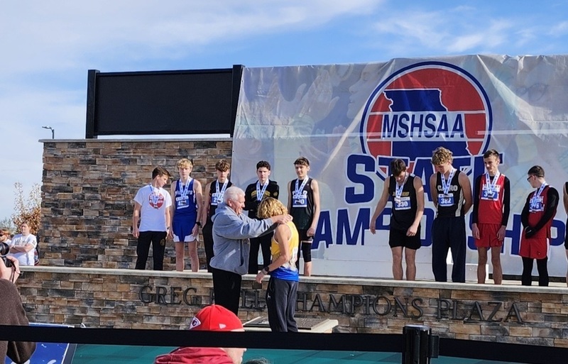 Coach Ratliff puts Carter's Medal around his neck at ceremony while 11 other runners look on in the background