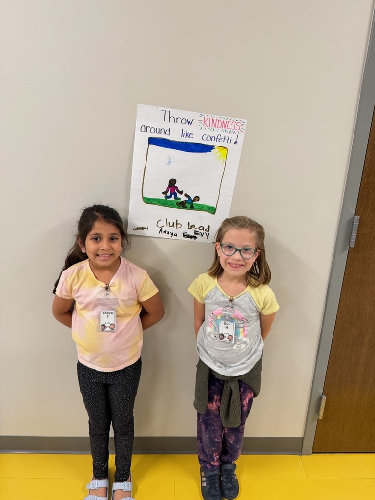 Two students smile at the camera with a poster on the wall  that reads "Throw Kindness around the confetti"