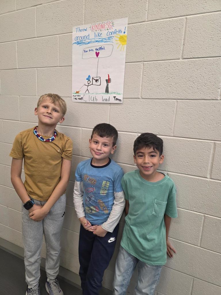 Three students smile at the camera with a poster on the wall  that reads "Throw Kindness around the confetti"