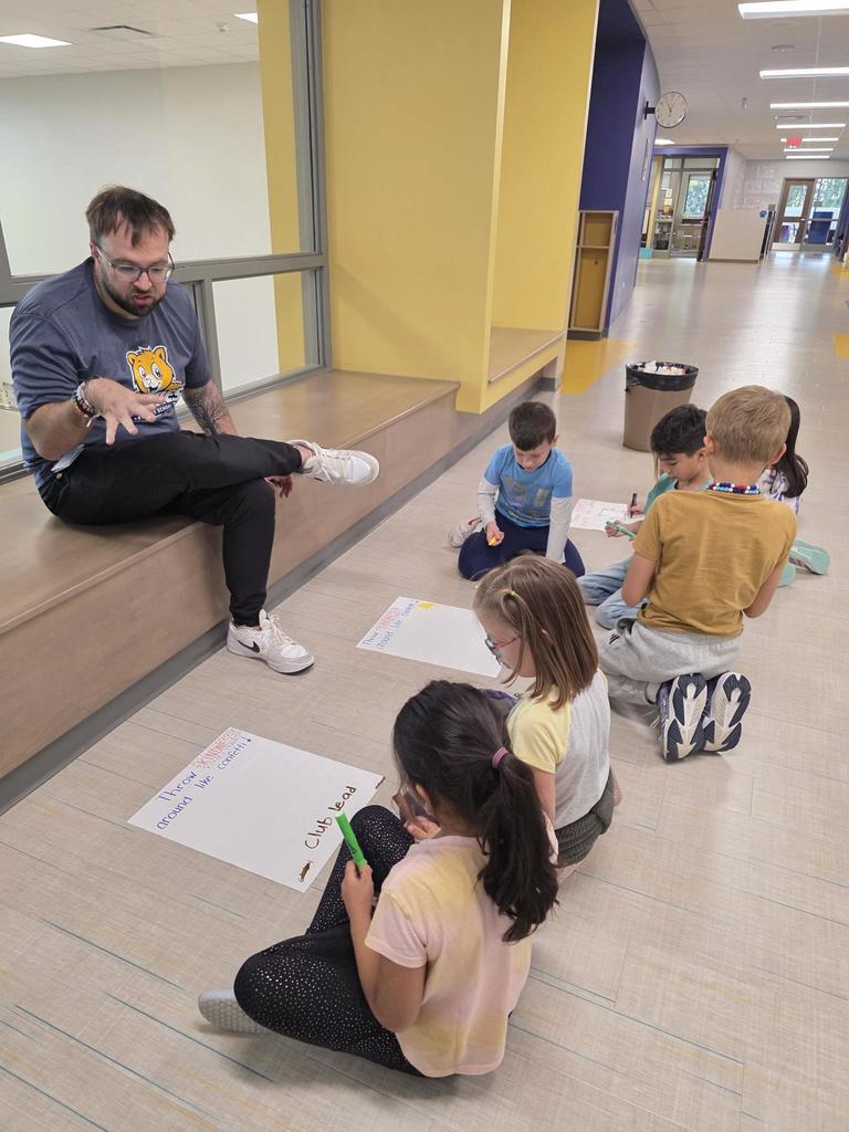 The teacher talks with students in a hallway as the students sit on the floor creating their posters