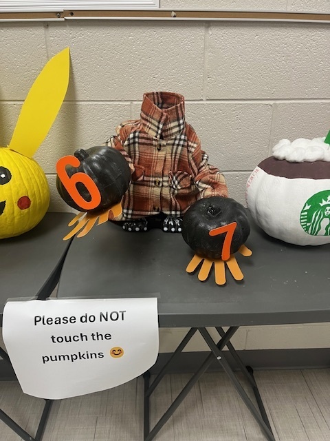 A pumpkin with a small flannel on decorated to look headless, holding two small pumpkins with the numbers 6 and 7
