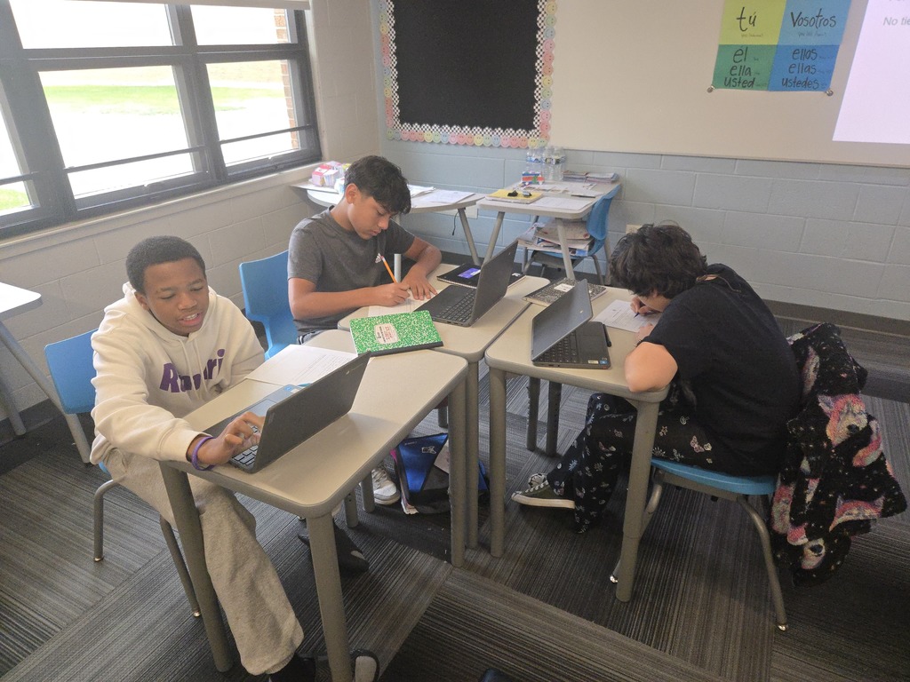 Three students sit at their desks writing and researching
