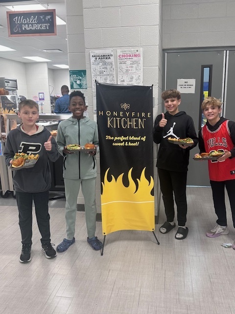 Students smile holding plates of food with a standing banner reading "Honeyfire Kitchen"