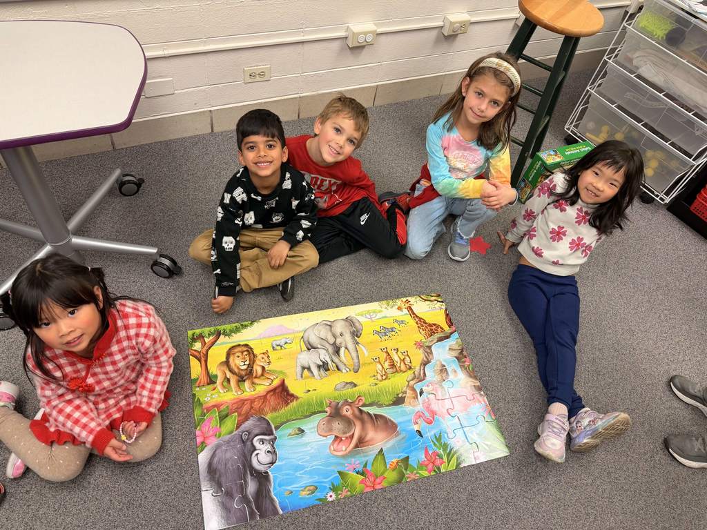 Students sit on the floor with an animal puzzle and smile at the camera