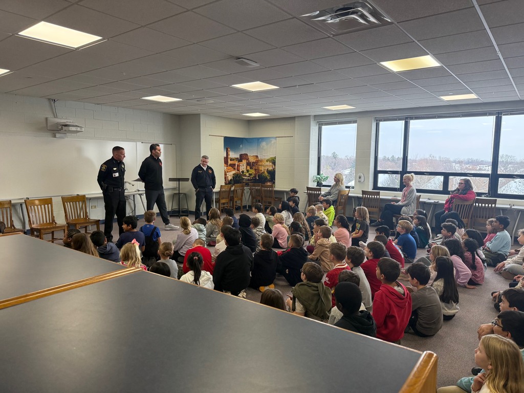 The visitors stand at the front of the classroom as the class sits on the floor and listens