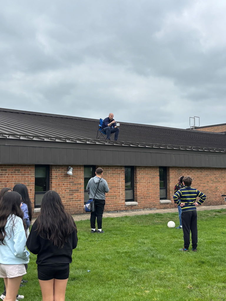 Principal on the roof eating lunch as students look on from the ground. 