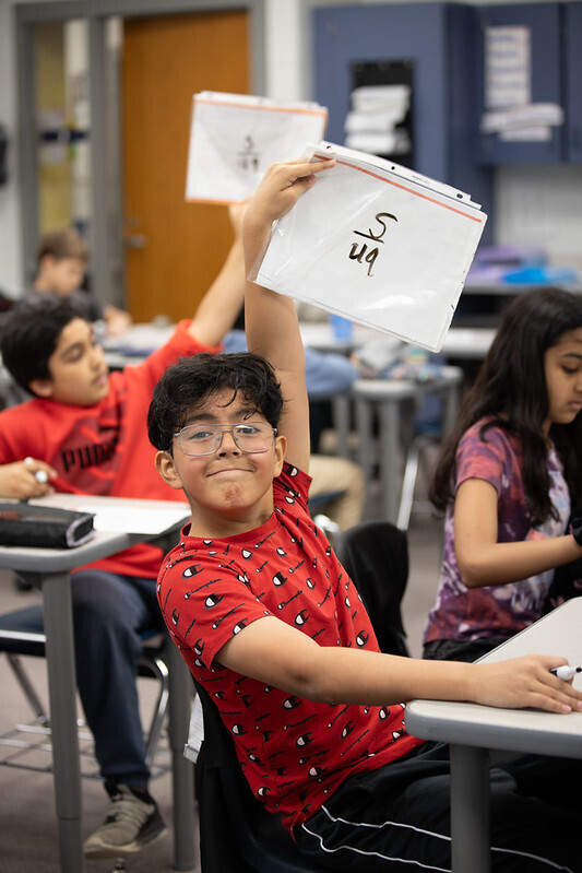 A student looks at the camera and raises his paper with a math answer written on it
