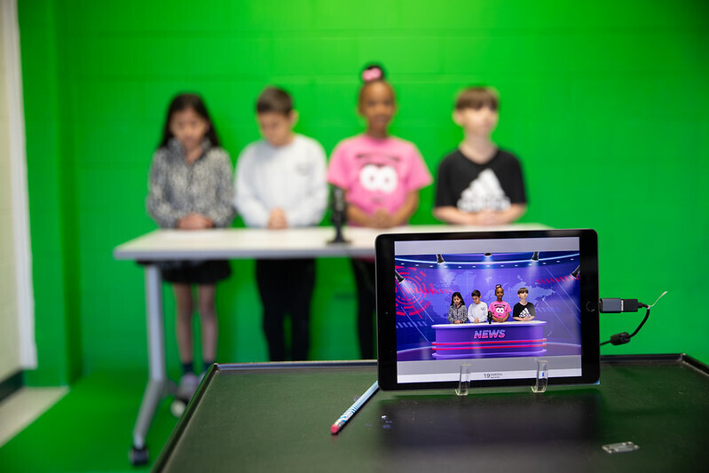 Four students stand in front of a green screen. A tablet records them. The tablet records them and shows the green screen at work, as the students look like they are standing at a news desk with a colorful background