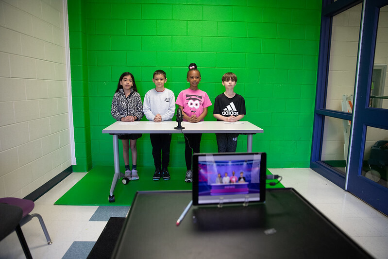 Four students stand in front of a green screen. A tablet records them. The students are in focus.