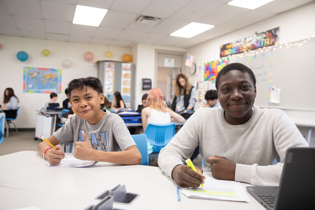 Two students sit together during class and smile at the camera. One student gives a thumbs up. 