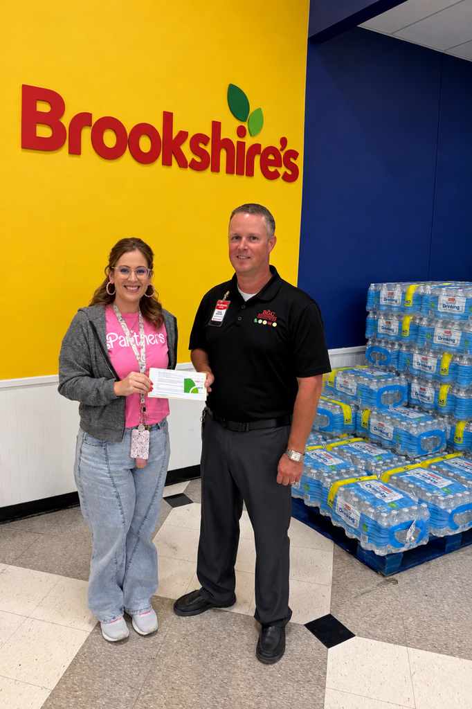 A woman in a pink shirt and a man in a black shirt stand before a wall with a Brookshire's logo. They hold a certificate and pose for a photo.
