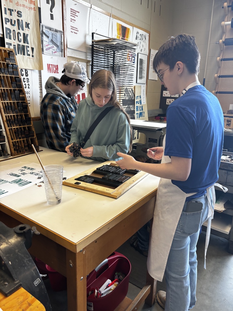 Three students work together at a printmaking station in an art classroom, arranging letterpress blocks on a tray while surrounded by posters, tools, and printing materials.