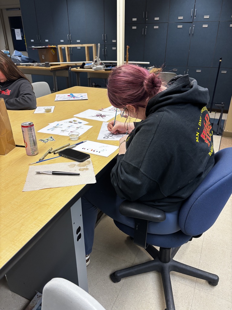 A student with pink hair tied in a bun sits at a table in a classroom, carefully painting detailed insect illustrations while art supplies, pens, and reference drawings are spread out around them.