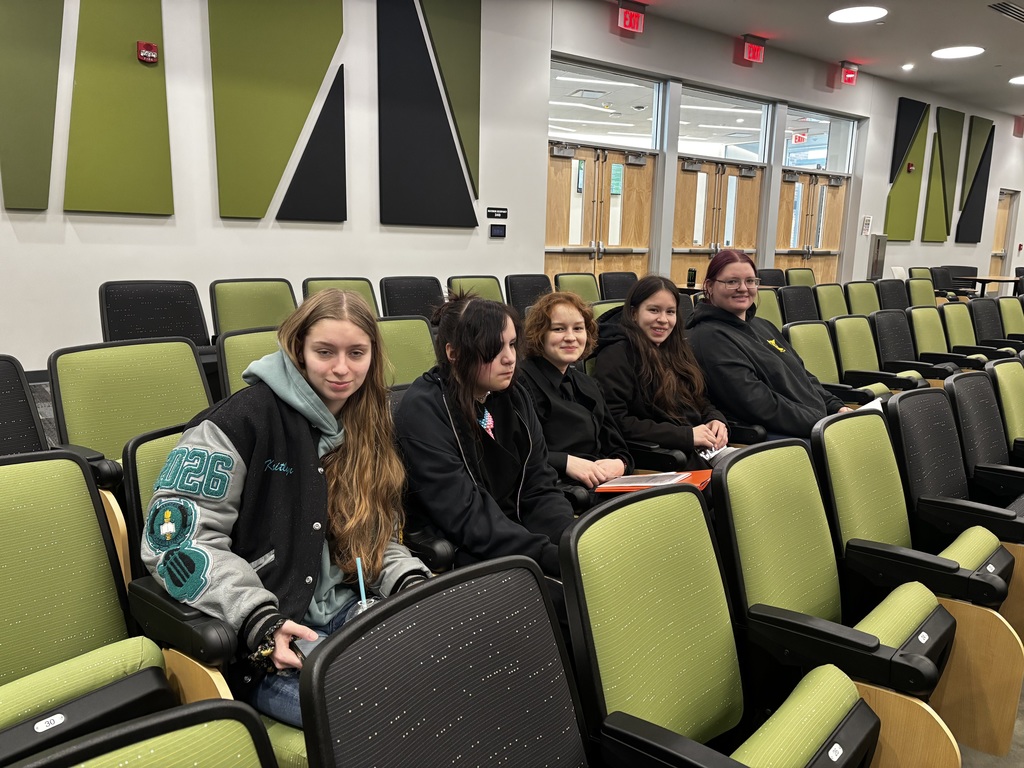 Five students sit together in a modern auditorium with green and black seats, looking toward the camera while waiting for an event or presentation.
