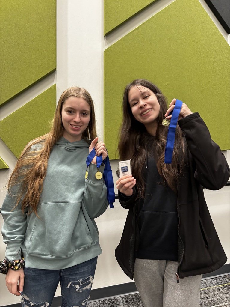 Two students stand indoors in front of green wall panels, smiling and holding up medals with blue ribbons, showing their awards.