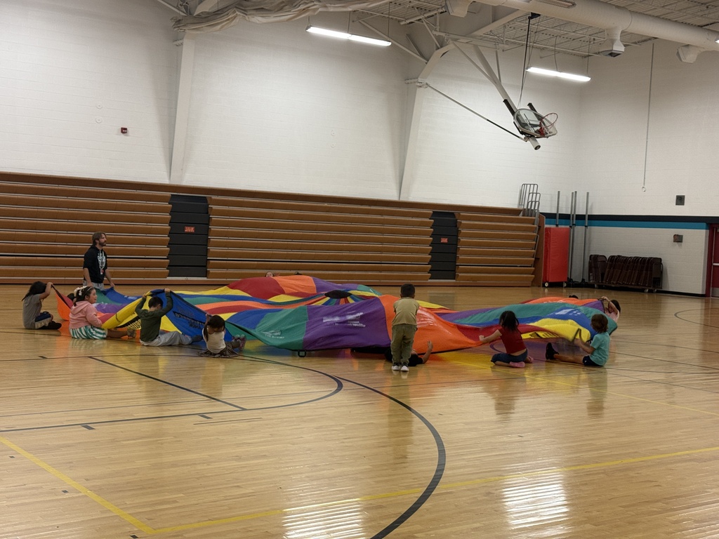 Students gather around a large, colorful rainbow parachute during a gym activity at Mahnomen Public School. Students are sitting, kneeling, and standing around the parachute while participating in the activity together.