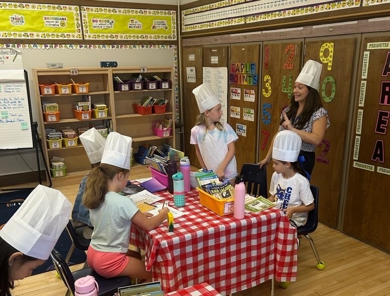 When third graders in Jena Amedeo’s class at Lakeview Elementary arrived at school recently, they were greeted with a special surprise – their classroom had been transformed into a “book tasting” café. Red and white gingham tablecloths covered the desks, place mats and chef hats were set out for each student, and baskets of books in a variety of genres were on the tables, ready to be “tasted.”  The special event served as the kickoff to the students’ first reading unit of the year, Building a Reading Life. To make the launch both fun and meaningful, the book tasting gave students a creative way to sample different genres of books they will encounter throughout the year during Readers Workshop.  “These are the kinds of experiences that stay with children,” Amedeo said. “My hope is that this book tasting will become a core memory for them, one that inspires a lifelong love of reading and reminds them that exploring books can be joyful and exciting.”  With their chef hats on, students browsed various books, spent time reading selections and recorded their impressions on “tasting sheets,” rating and reflecting on each new title they explored. Students laughed, shared discoveries and proudly announced their new favorites. At one point, a student shouted out, “This is the best day ever!” a sign of the enthusiasm in the room. The activity encouraged students not only to try something new, but also to think about what kinds of stories spark their interest and excitement.  The event was a hit with students and staff alike, serving as both a celebration of reading and an invitation to begin a yearlong journey into new genres, stories and adventures.