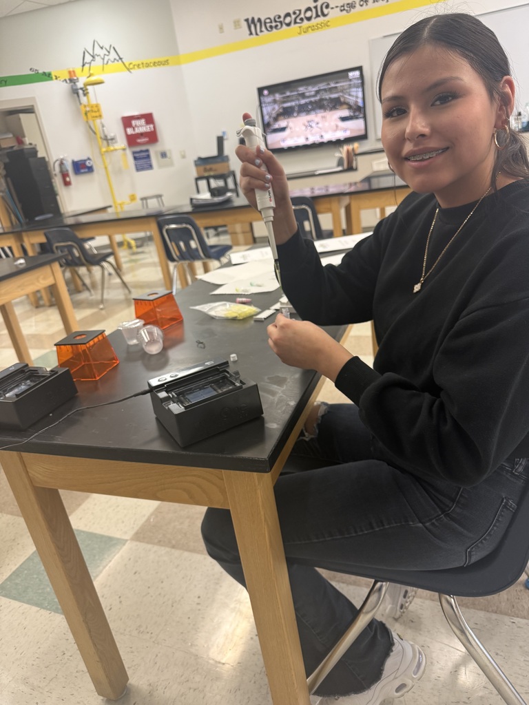 A smiling female student is sitting at a lab table conducting a gel electrophoresis experiment. In the background, the District basketball games are playing on the Smartboard.