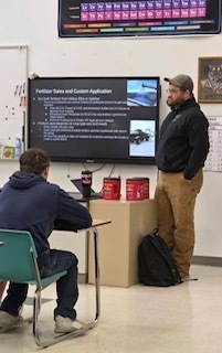 An instructor stands at the front of a classroom next to a large screen displaying a presentation about fertilizer safety or application. A student sits at a desk facing the screen, listening. The room includes classroom posters, a periodic table chart on the wall, and typical school furniture like desks and chairs.