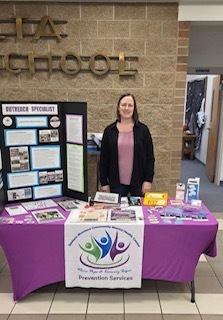 A woman stands behind an informational table set up indoors, likely in a school lobby. The table is covered with a purple cloth and displays brochures, pamphlets, and a tri-fold presentation board labeled “Outreach Specialist.” A banner on the front reads “Prevention Services” with a colorful logo. A wall behind her includes large lettering that appears to say “High School,” and a hallway is visible to the side.