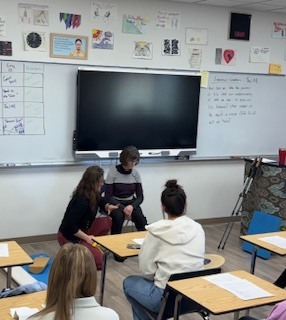 A small group of students sits in a classroom gathered around a desk at the front of the room. Two students sit close together while another leans in, appearing to talk or check something on a phone or paper. Other students sit at desks nearby. The whiteboard behind them has notes and drawings, and a large screen is mounted at the center.