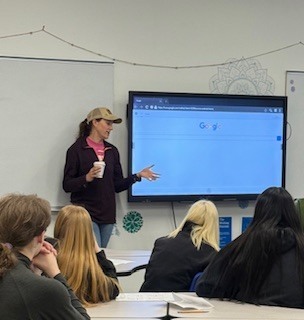 A teacher stands at the front of a classroom holding a drink and gesturing while speaking to students seated at desks. Behind the teacher is a large interactive screen displaying a Google homepage, with a whiteboard and classroom decorations visible on the wall.