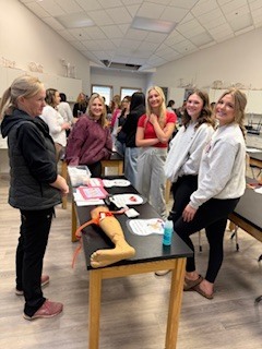 A group of smiling students stand around a classroom table during a hands-on activity. On the table are materials including a model of a leg, papers, and tools. An instructor stands to the side engaging with the group. The classroom has lab tables, stools, and bright overhead lighting, suggesting a science or health-related lesson.