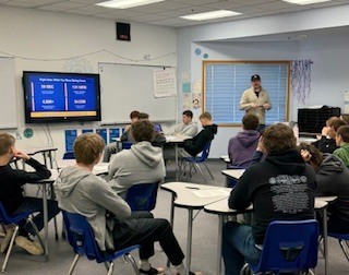 A classroom of students sits at individual desks arranged in groups while a teacher stands at the front near a large screen displaying a quiz or review game. The students face forward, some leaning back in their chairs, as the teacher appears to lead the activity.