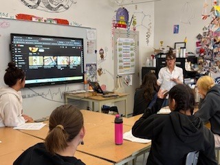 A female teacher sits at the front of a classroom speaking to a small group of students gathered around tables. An interactive screen behind the teacher displays a video interface, while students sit with notebooks and water bottles, listening and facing the front.