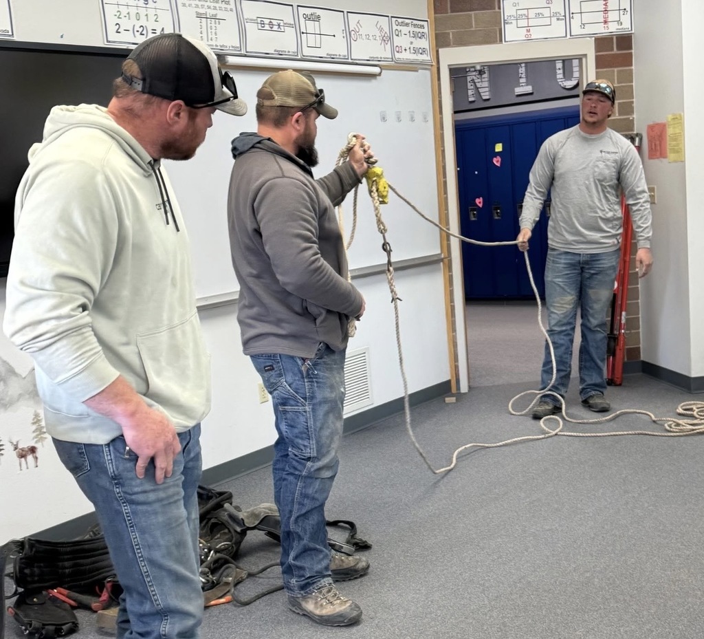 Three men stand in a classroom-like setting, demonstrating rope handling techniques. One man holds a coiled rope with a metal hook while another stands across the room holding the other end, and a third observes nearby. Posters with math diagrams are visible on the wall, and equipment such as harnesses lies on the floor.