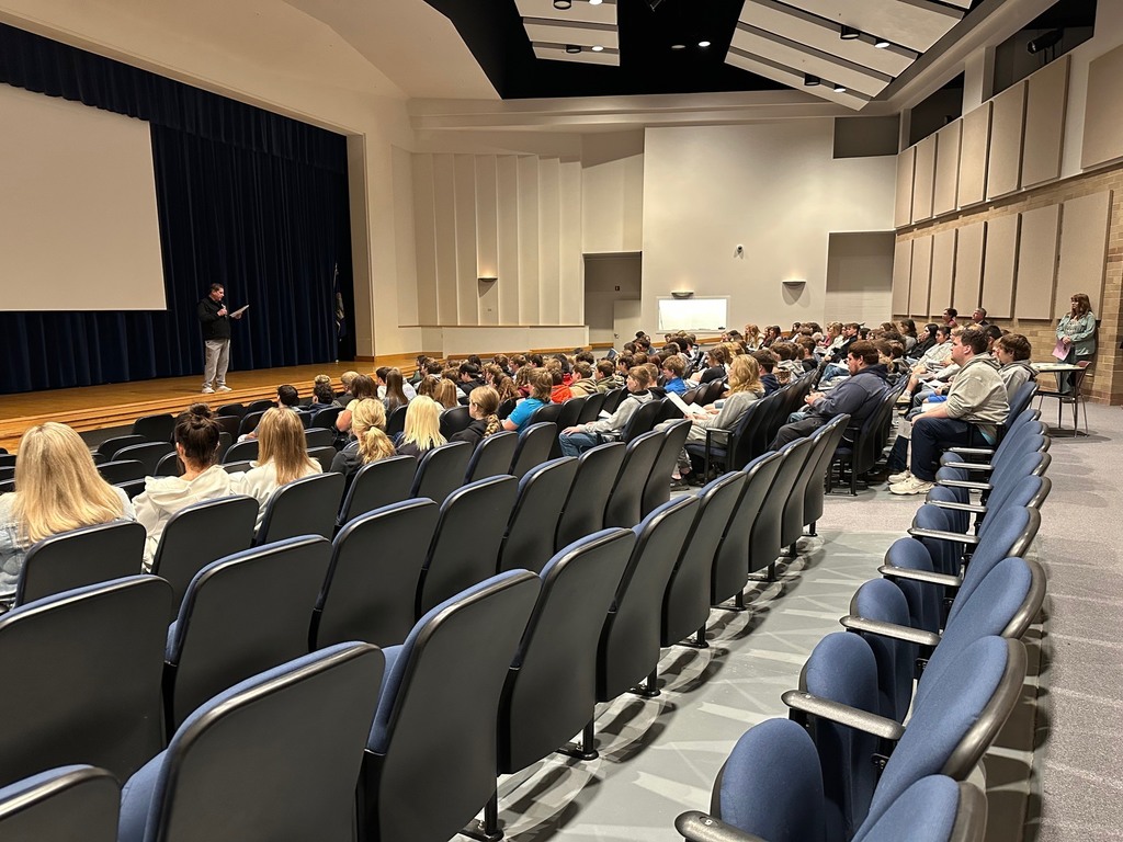A large school auditorium filled with students seated in rows of chairs, facing a stage where a person stands speaking and reading from a paper. The room has high ceilings with acoustic panels, a projection screen on the stage, and a few adults standing along the side observing.