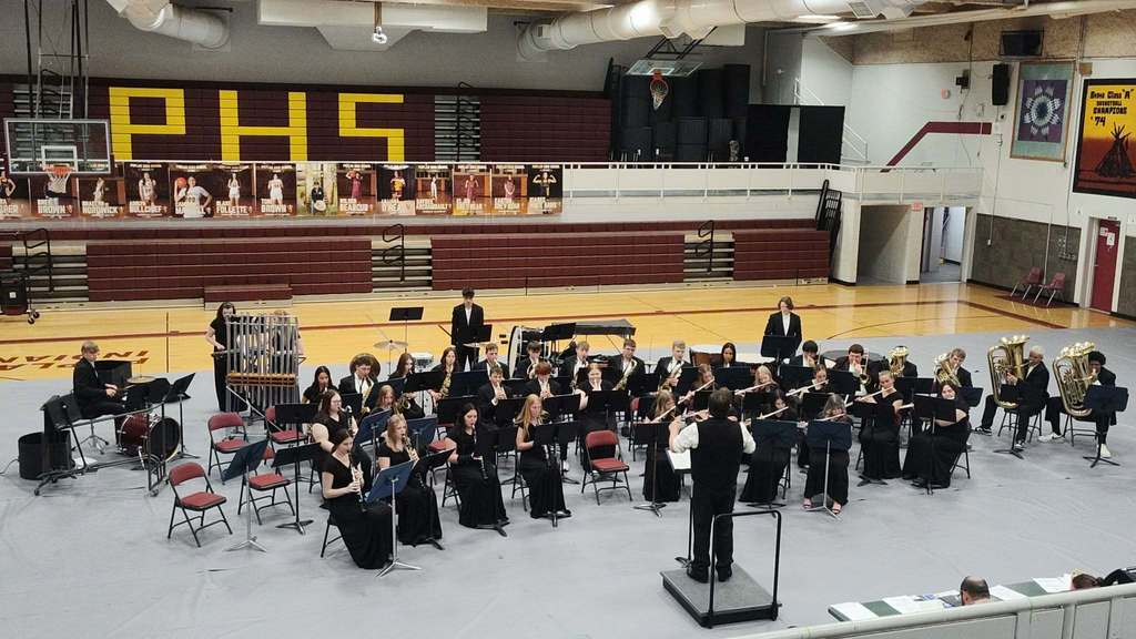 High school concert band performing in a gymnasium, with students seated in semicircles holding wind and brass instruments while a conductor stands on a podium directing in the foreground; percussion instruments are set up on the left, and bleachers and school banners are visible in the background.