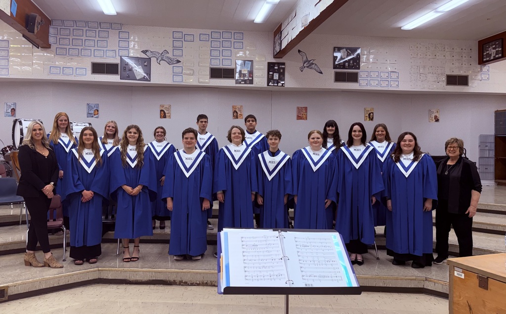High school choir group standing in two rows inside a music classroom, wearing matching blue robes with white stoles labeled “MHS,” posing for a formal group photo with directors on either end; a music stand with open sheet music is visible in the foreground and classroom walls display music-related posters and achievements in the background.