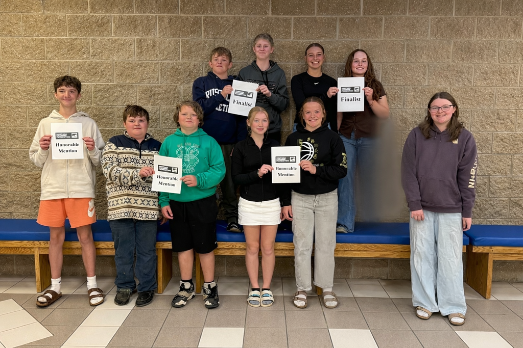 Alt text: A group of middle school students stand in two rows against a tan brick wall, smiling and holding certificates. Some students hold signs that read “Finalist,” while others hold “Honorable Mention.” They are dressed casually in hoodies, sweatshirts, shorts, and jeans. The students in the back row stand on a bench, while those in front stand on the floor. 