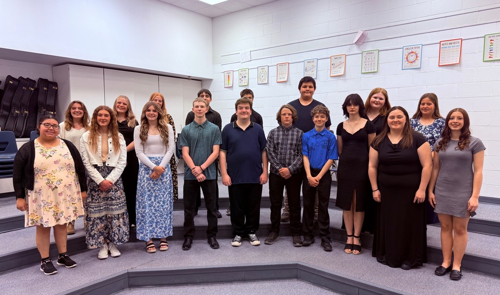 A group of approximately 16 middle or high school choir students pose together on tiered risers inside a music classroom. The students are dressed in semi-formal concert attire, including dresses, skirts, polos, and dress shirts in neutral and dark colors. Music theory and choir posters hang along the back wall, and instrument cases are stacked on the left side of the room. The students are smiling and standing in rows for a formal choir group photo.