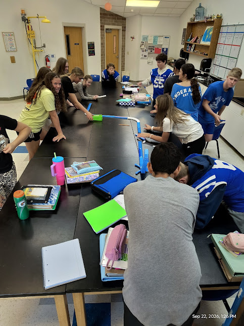 A group of students gathered around a cluster of desks building a large hotwheels track.