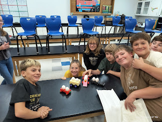 A group of students posing by their egg cars. On the left is a red car with a small Christmas tree on the front, then a yellow car with the word "Butter" in blue on the side, a pink car with a jack-o-lantern on it, and finally a black car with a broken pirate ship mast on it. 