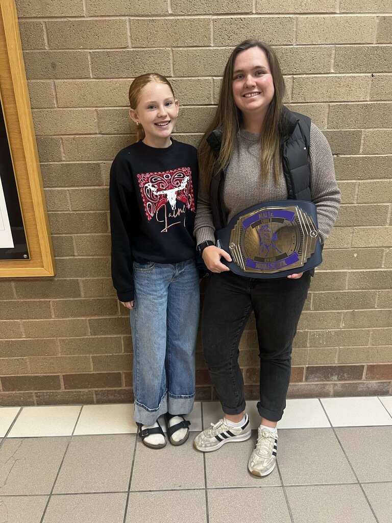 Haidyn H., a young student with red hair pulled back, stands beside her teacher, Mrs. Paul, smiling at the camera. Haidyn is wearing a black sweatshirt with a red-and-white Western design, cuffed blue jeans, and black sandals. Mrs. Paul has long brown hair and is wearing a gray sweater, black quilted vest, black pants, and white sneakers. Mrs. Paul is holding a large purple-and-gold championship-style belt recognizing the Malta Middle School Core 3 Award. They are standing in front of a tan brick wall with a tiled floor.