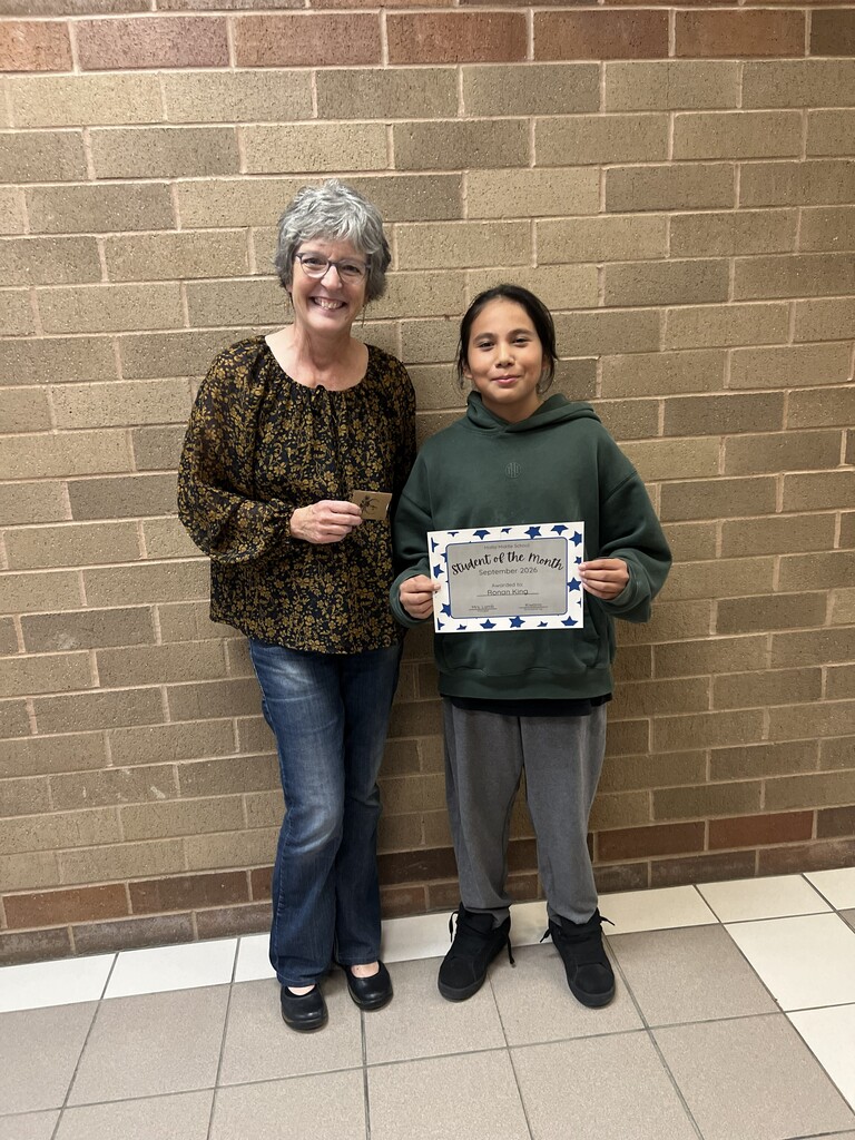 Adult Mardy Demaris and student Ronan stand side by side in front of a tan brick wall at Malta Middle School. Mardy, wearing glasses, a black blouse with a gold floral pattern, and blue jeans, smiles while holding a small card. Ronan wears a dark green hoodie, gray sweatpants, and black shoes. He holds a certificate recognizing him as the September 2026 Student of the Month. Both face the camera and smile.