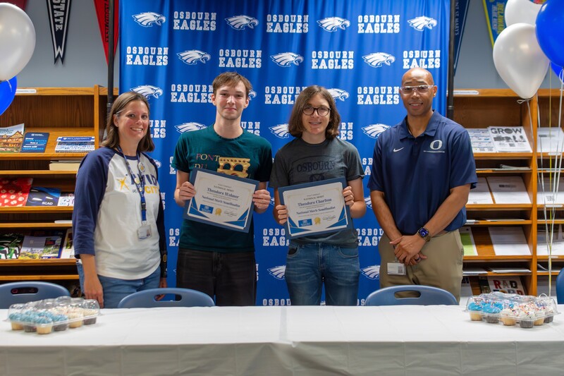 Two students holding up certificates in front of backdrop in between Principal Dr. Jennifer Chapman and Superintendent Dr. Kevin Newman.