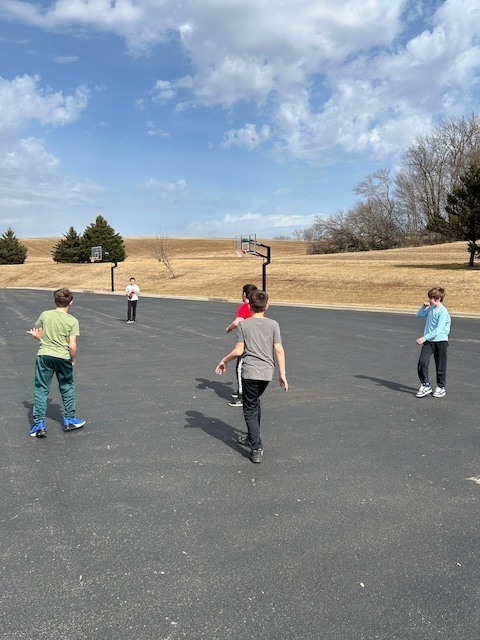 5th graders outside on blacktop playing football grass trees and basketball hoops in the background