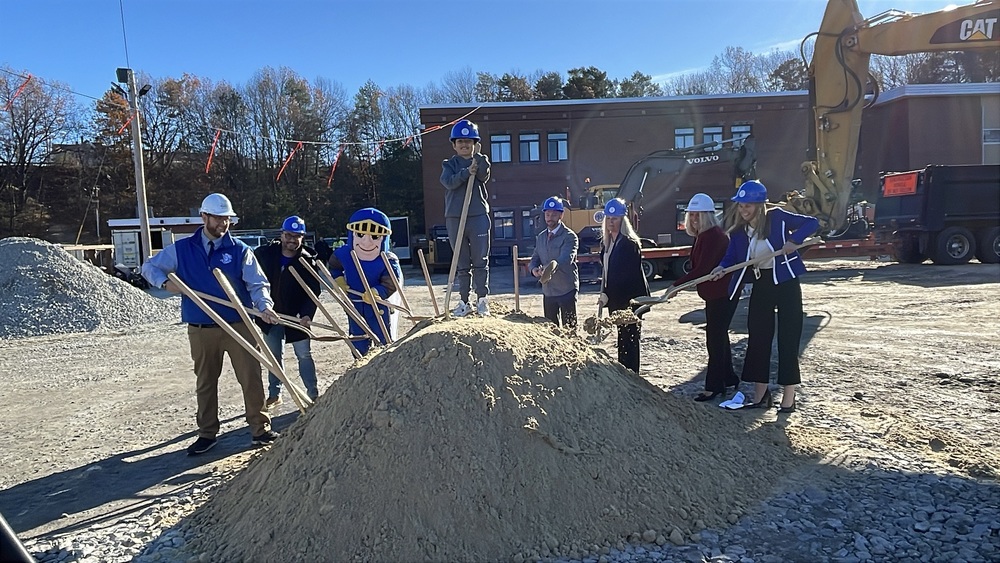 School district leaders and the Parkside mascot dig shovels into a big pile of dirt on a sunny November day while wearing white and blue hardhats. A young student stands atop the pile of dirt with a shovel and hardhat