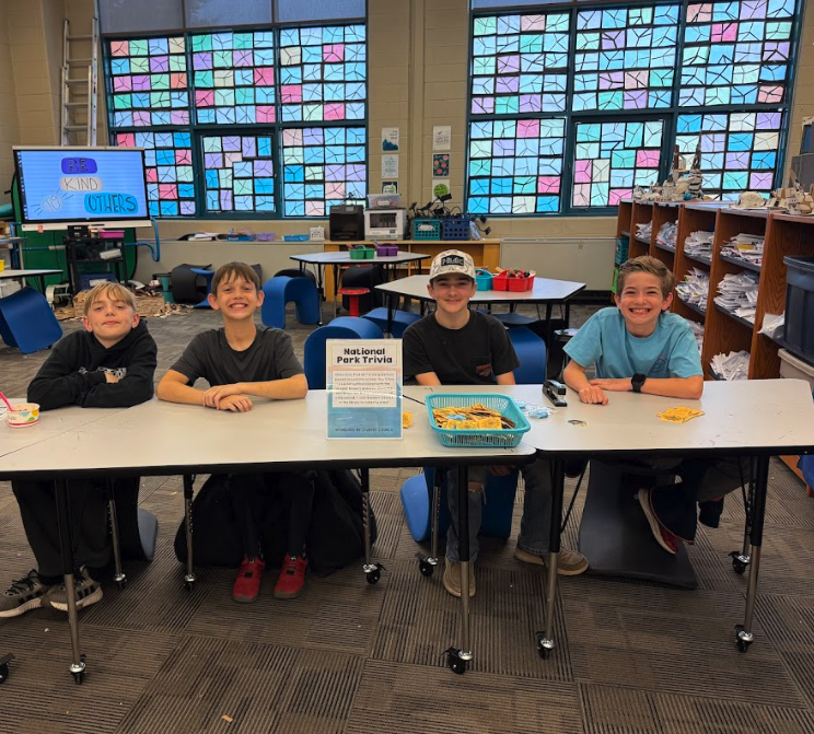 Four boys in a classroom with a table and chairs. A sign is on the table, and they are all looking at the camera.