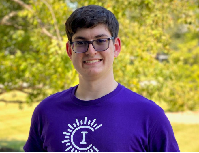 A person in a purple t-shirt with a white logo smiles. They wear glasses and stand in front of leafy trees.