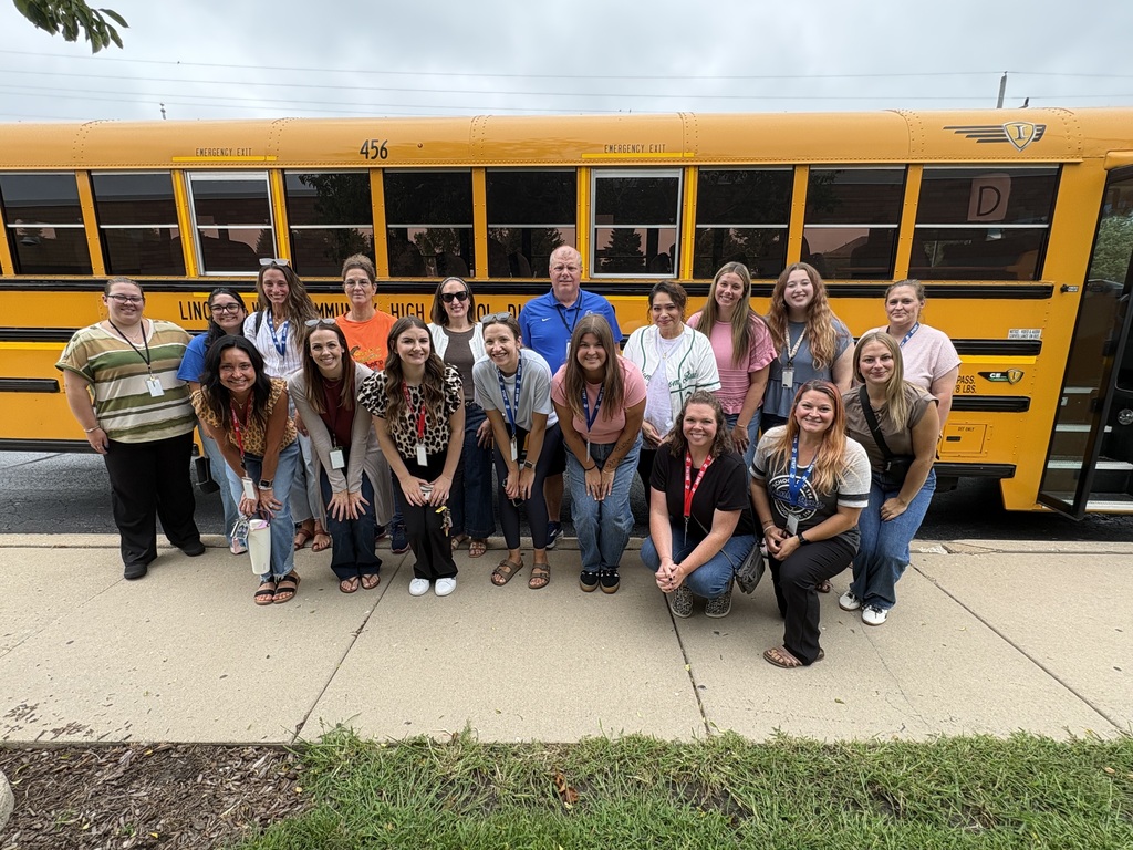 Group of adults and children in front of a yellow school bus on a sidewalk.
