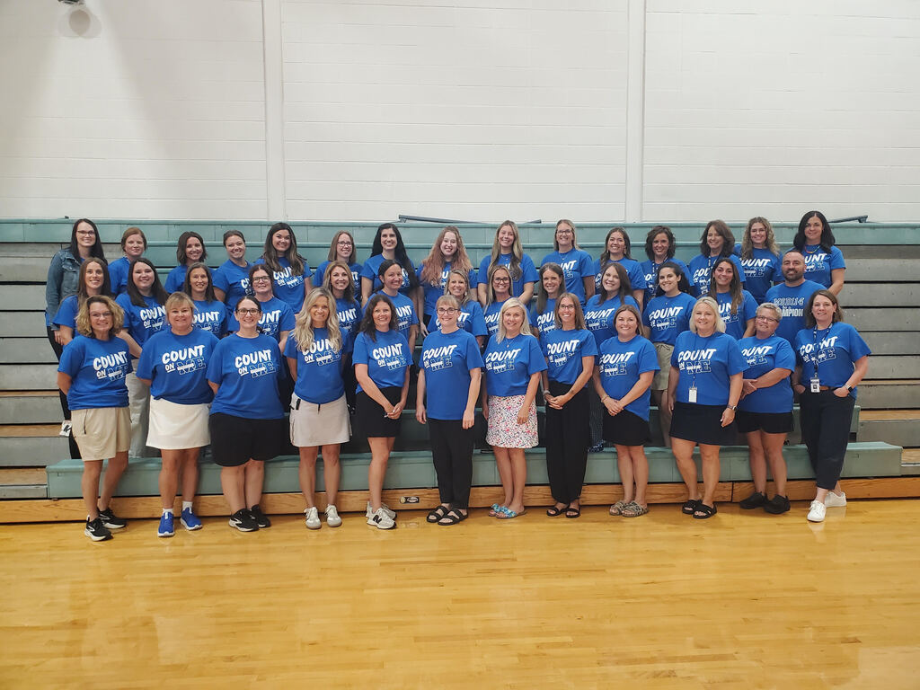 A group of women in blue shirts, some with white text, pose for a photo in a gym with wooden floor and bleachers.