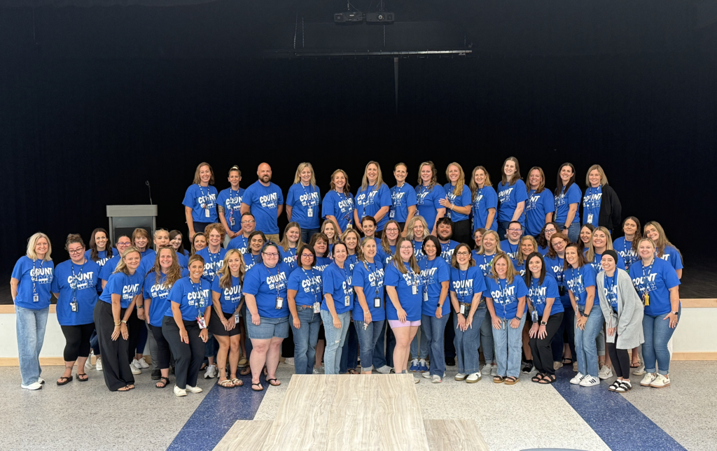 A group of people in blue shirts, some with "WELCOME" on them, pose for a photo on a stage.