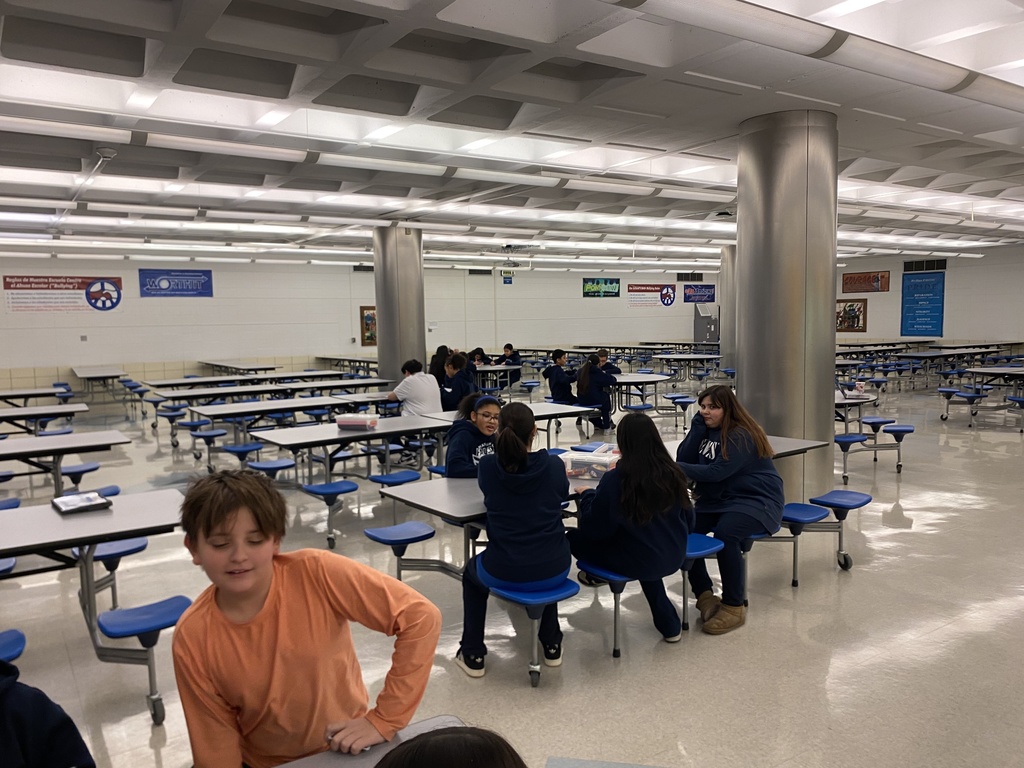 A photo of the spacious cafeteria at Mannheim Middle School, where students are seated in small groups at round tables with blue stools. The room features high ceilings with bright lighting and large silver structural columns. In the foreground, a student in an orange long-sleeved shirt leans into the frame, while other students converse and relax in the background.