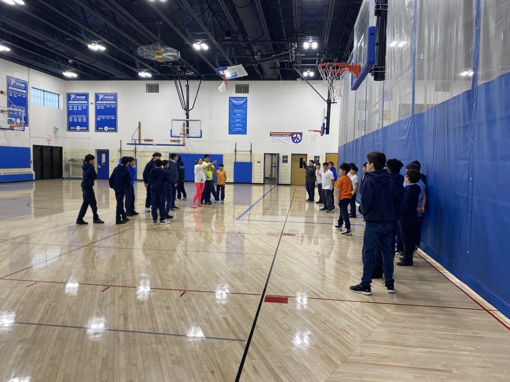 A photo of a physical education class taking place inside the Mannheim Middle School gymnasium. A teacher is actively instructing a group of students who are gathered on the wooden basketball court and lined up along a large blue divider wall. The bright facility features basketball hoops and school banners hanging in the background.