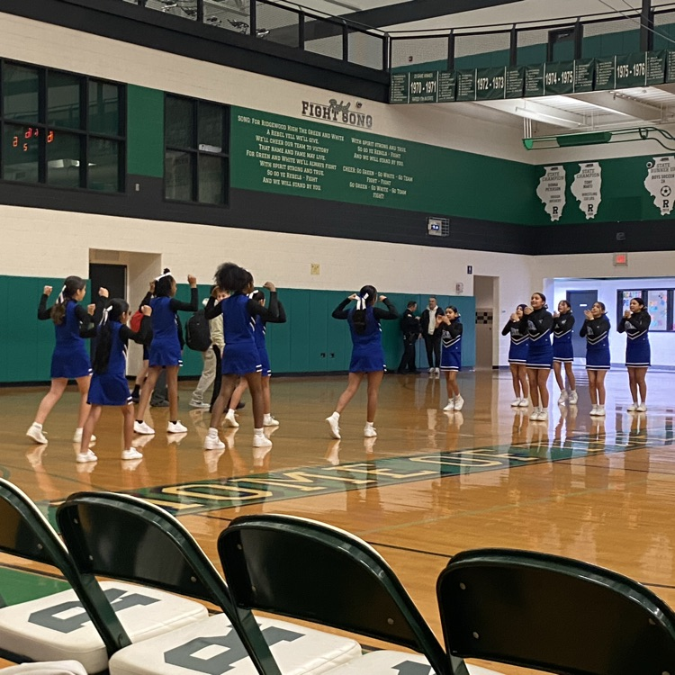 A photo of the Mannheim School District cheerleading squad performing a routine on the wooden basketball court at Ridgewood High School. The cheerleaders, dressed in blue and white uniforms, are arranged in two lines with their backs to the camera, raising their arms. In the background, a green wall displays the school's "Fight Song," and spectators watch from the side.